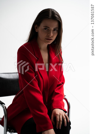 Woman in a red blazer sitting on a chair, showcasing professional confidence and modern fashion style in a studio portrait, looking seriously at the camera 137081587