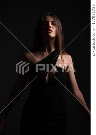 Young woman posing in a studio with dramatic lighting, showcasing a stylish black dress and creating an mood of elegance and allure, communicating fashion, femininity, and identity Young woman posing in a studio with dramatic lighting, showcasing a stylish black dress and creating an mood of elegance and allure, communicating fashion, femininity, and identity 137081789