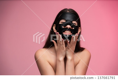 Happy young woman enjoying a skincare ritual, applying a purifying charcoal face mask, smiling on a vibrant pink background, promoting self-care and beauty Happy young woman enjoying a skincare ritual, applying a purifying charcoal face mask, smiling on a vibrant pink background, promoting self-care and beauty 137081791