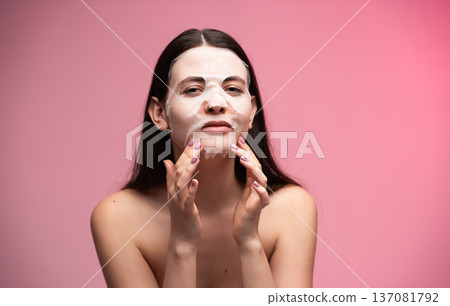 Young woman applying a hydrating sheet facial mask, performing a modern self-care skincare routine for beauty and wellness on a pink studio background 137081792