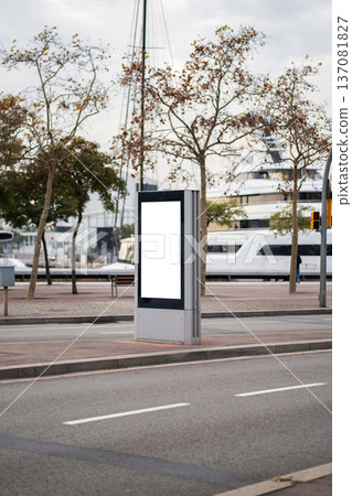 Blank vertical digital screen display mockup positioned in a modern urban street with trees and large luxury yachts moored at a harbor in the background 137081827