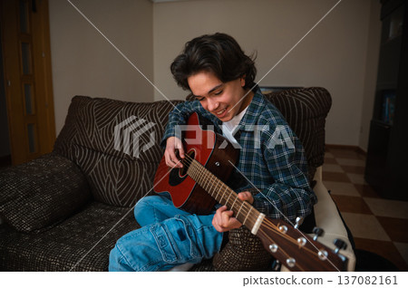 Teenager Boy Playing Acoustic Guitar At Home On Cozy Couch While Smiling And Strumming Teenager Boy Playing Acoustic Guitar At Home On Cozy Couch While Smiling And Strumming 137082161