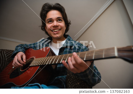 Teenage Boy Playing Acoustic Guitar At Home Smiling While Strumming In Cozy Casual Setting Teenage Boy Playing Acoustic Guitar At Home Smiling While Strumming In Cozy Casual Setting 137082163
