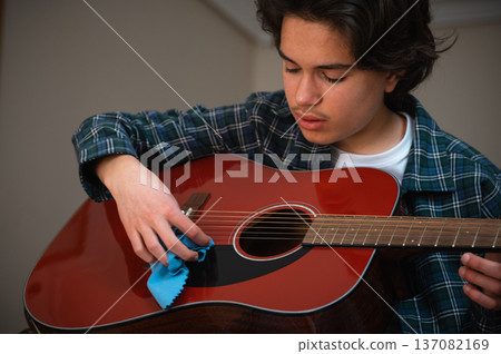 Teenage Boy Cleaning and Playing Red Acoustic Guitar in Cozy Home Interior Teenage Boy Cleaning and Playing Red Acoustic Guitar in Cozy Home Interior 137082169