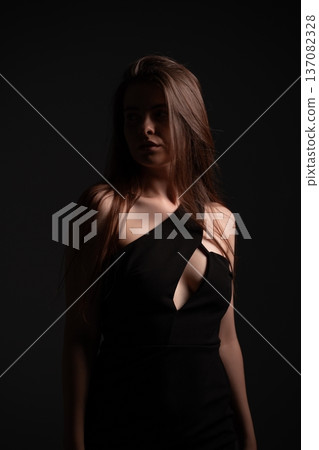 Young woman posing in a black dress with an asymmetrical neckline and cutout, illuminated by dramatic lighting against a dark background, conveying a sense of elegance, intrigue, and mystery 137082328