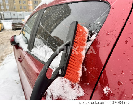 a girl cleans her personal car from snow with a black-and-orange car brush in the courtyard of an apartment building 137084562