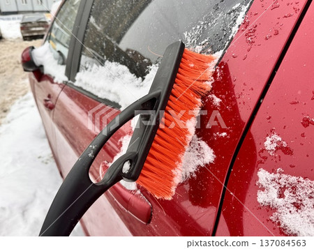 a girl cleans her personal car from snow with a black-and-orange car brush in the courtyard of an apartment building 137084563