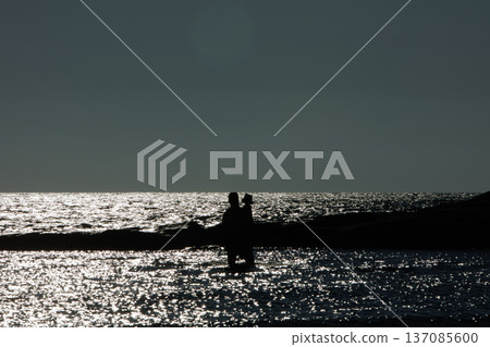 Silhouette of an adult with a child standing in shallow water at sunset in Vlora, Albania, captured on August 25, 2025. The sparkling sea reflects the fading light Silhouette of an adult with a child standing in shallow water at sunset in Vlora, Albania, captured on August 25, 2025. The sparkling sea reflects the fading light 137085600