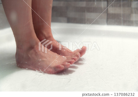 Close-up of Female bare feet standing on wet shower floor with water droplets falling from above, tiled wall visible in the background Close-up of Female bare feet standing on wet shower floor with water droplets falling from above, tiled wall visible in the background 137086232