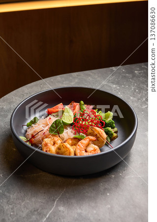 Delicious shrimp with fresh lime, vibrant vegetables, rice noodles served in a stylish black bowl on grey stone table, Asian cuisine concept Delicious shrimp with fresh lime, vibrant vegetables, rice noodles served in a stylish black bowl on grey stone table, Asian cuisine concept 137086630