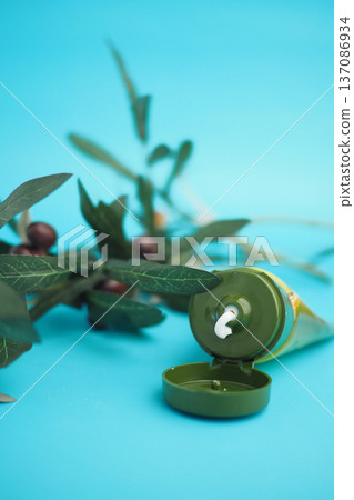 A tube of olive cream is placed next to an olive branch on a blue surface A tube of olive cream is placed next to an olive branch on a blue surface 137086934