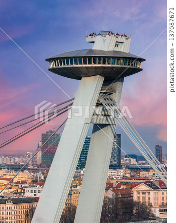 Close up shows the UFO observation deck and tilted pylon of SNP Bridge in Bratislava, Slovakia, at sunset, with radiating cables, city rooftops, and tiny figures visible. 137087671