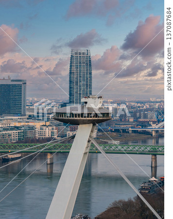 Aerial view of Bratislava, Slovakia, shows the UFO deck on SNP Bridge over the Danube, a modern skyline, a high rise tower, green steel bridges, and a pastel sunset sky. Aerial view of Bratislava, Slovakia, shows the UFO deck on SNP Bridge over the Danube, a modern skyline, a high rise tower, green steel bridges, and a pastel sunset sky. 137087684