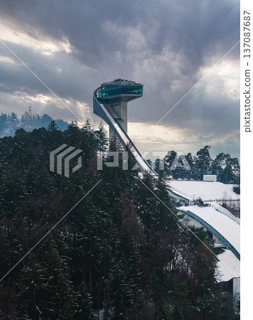 Aerial view of Bergisel Ski Jump in Innsbruck, Austria, angular tower and glass platform rise over snowy trees, curving inrun and icy track stand out in late afternoon light. 137087687