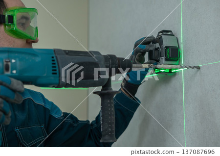 A man measures distances with a laser level and drills into a wall with a hammer drill. A man measures distances with a laser level and drills into a wall with a hammer drill. 137087696