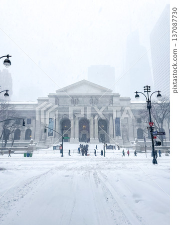 A heavy snowstorm covers the New York Public Library main branch, with lion statues, grand columns, and pedestrians crossing toward glowing lights in Midtown. A heavy snowstorm covers the New York Public Library main branch, with lion statues, grand columns, and pedestrians crossing toward glowing lights in Midtown. 137087730