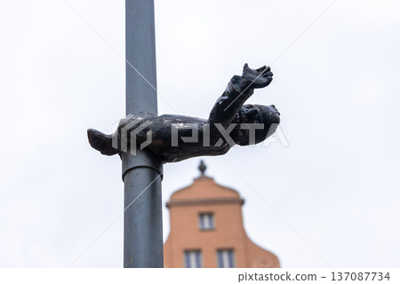 Small bronze dwarf clings horizontally to a gray pole, arms outstretched. A peach tenement with a stepped gable rises behind. Wroclaw, Poland, muted daylight. Small bronze dwarf clings horizontally to a gray pole, arms outstretched. A peach tenement with a stepped gable rises behind. Wroclaw, Poland, muted daylight. 137087734
