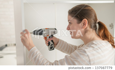 A Caucasian woman uses a screwdriver to tighten a screw while assembling a cabinet. 137087779
