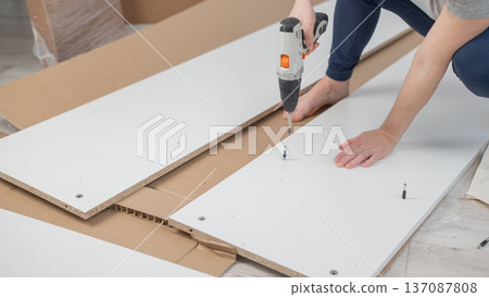 A Caucasian woman uses a screwdriver to tighten a screw while assembling a cabinet. A Caucasian woman uses a screwdriver to tighten a screw while assembling a cabinet. 137087808
