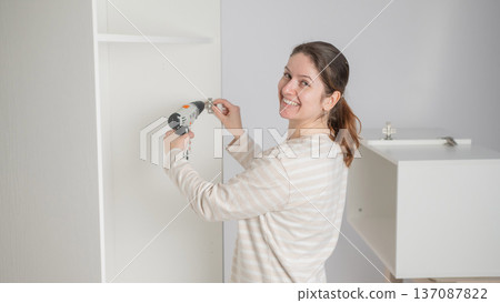 A Caucasian woman uses a screwdriver to tighten a screw while assembling a cabinet. 137087822