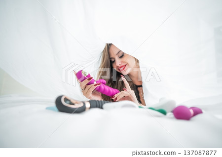 Woman in lace lingerie holds a dildo. A beautiful Caucasian woman lies under the sheets and holds a dildo.  137087877