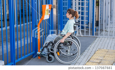 A woman in a wheelchair uses a touch-sensitive Braille help button.. A woman in a wheelchair uses a touch-sensitive Braille help button.. 137087881