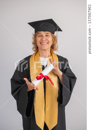 Happy senior woman in graduation gown holding diploma on white background.  137087901
