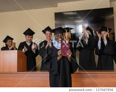Happy African woman in hijab receiving diploma and classmates applauding her. Happy African woman in hijab receiving diploma and classmates applauding her. 137087956