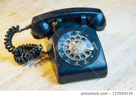 A black rotary phone with a coiled cord rests on a wooden table. The setting shows a simple room with natural light coming through a nearby window. A black rotary phone with a coiled cord rests on a wooden table. The setting shows a simple room with natural light coming through a nearby window. 137089134
