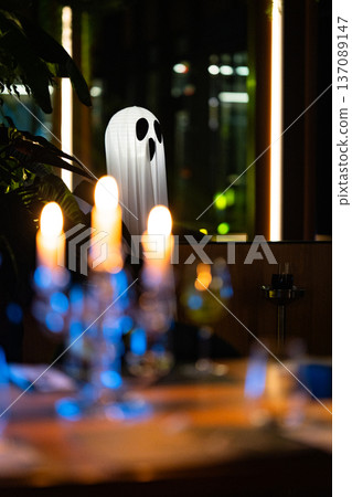 A ghost figure made from white fabric sits quietly at a table in a restaurant. Candles flicker softly, casting warm light. The atmosphere is dark but inviting, with subtle colors in the background. 137089147
