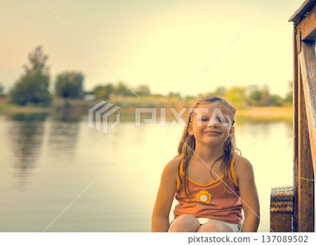 A cute little girl sits on a wooden pier at the shore of a lake. The photo was taken during the golden hour, at sunset. It represents childhood, summer vacations, looking toward the future, and A cute little girl sits on a wooden pier at the shore of a lake. The photo was taken during the golden hour, at sunset. It represents childhood, summer vacations, looking toward the future, and 137089502