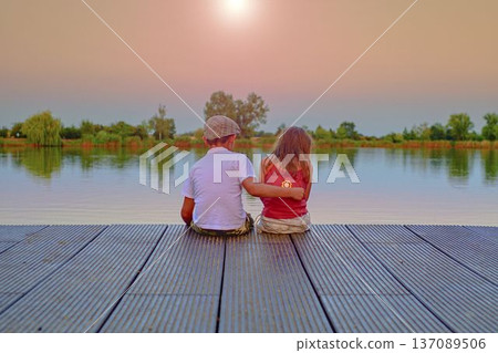 A little boy wearing a flat cap and a cute young girl sit together on a wooden pier by the lake. Small children sit on the pier at the side of a pond. The concept of a carefree childhood and summer A little boy wearing a flat cap and a cute young girl sit together on a wooden pier by the lake. Small children sit on the pier at the side of a pond. The concept of a carefree childhood and summer 137089506