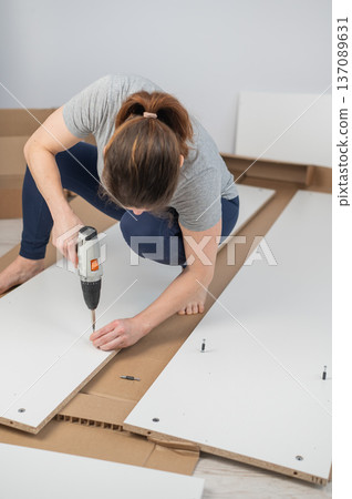 A Caucasian woman uses a screwdriver to tighten a screw while assembling a cabinet. A Caucasian woman uses a screwdriver to tighten a screw while assembling a cabinet. 137089631