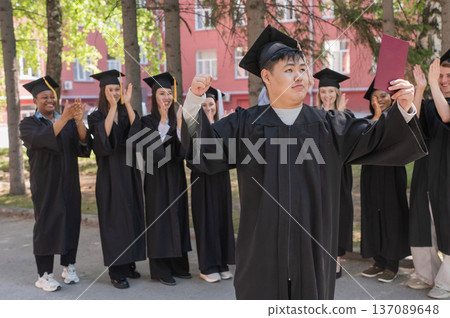 Young Asian man holds diploma while classmates applaud her.  137089648