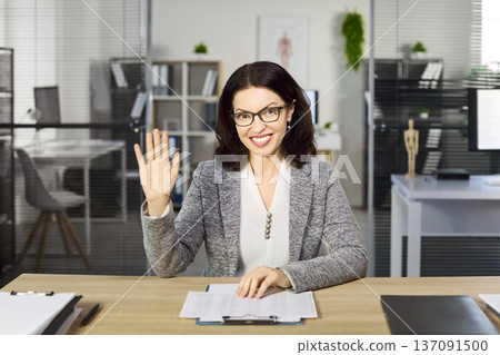 Friendly businesswoman waving hello in office during online meeting or webinar Friendly businesswoman waving hello in office during online meeting or webinar 137091500