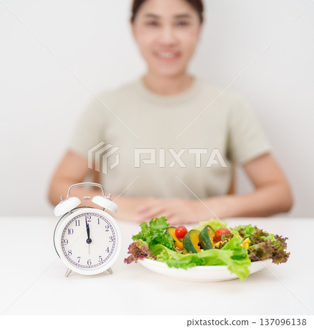 happy smiling woman with fresh organic vegetables plate and alarm clock. intermittent fasting, IF, meal time, nutrition, weight loss, clean eating, Healthy food eating, Wellness, dieting and lifestyle 137096138