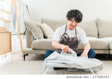 A man in an apron ironing a shirt in the living room 137098167