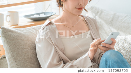 一位亞洲女性坐在客廳的沙發上,看著她的智慧型手機。 一位亞洲女性坐在客廳的沙發上,看著她的智慧型手機。 137098703