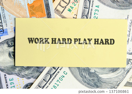 A bright yellow note, inscribed with Work Hard Play Hard, is gently positioned on a bed of scattered US dollar bills 137100861