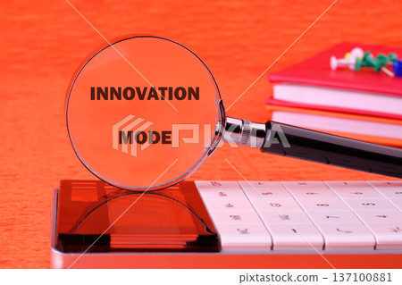 A magnifying glass highlights the words Innovation Mode on a calculator, accompanied by a stack of books and colorful pushpins on an orange desk A magnifying glass highlights the words Innovation Mode on a calculator, accompanied by a stack of books and colorful pushpins on an orange desk 137100881
