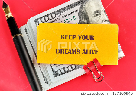A vibrant red surface displays a stack of US hundred dollar bills, a sleek fountain pen, and a yellow card urging Keep Your Dreams Alive A vibrant red surface displays a stack of US hundred dollar bills, a sleek fountain pen, and a yellow card urging Keep Your Dreams Alive 137100949
