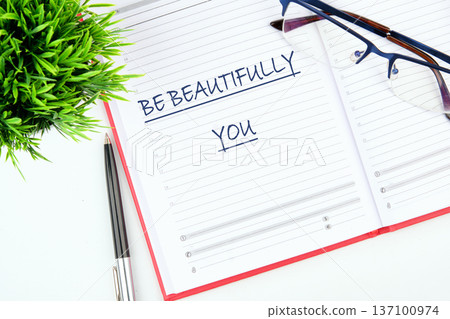 Uplifting words Be Beautifully You written in a diary. A refined pen, elegant glasses, and a vibrant green plant adorn a clean, sunlit desk setting 137100974