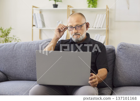 Mature man adjusts glasses while working on laptop at home living room interior 137101168