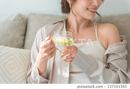 A woman sitting on a sofa drinking lemon water, detox water A woman sitting on a sofa drinking lemon water, detox water 137101563