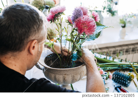 Man arranges pink and purple asters in vintage white urn using wire mesh. dopamine decor flowers, bright floral energy, mood boosting bouquets, colorful flower therapy 137102805