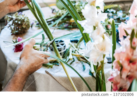 Man cuts gladiolus stem with green pruning shears on table. Artisan at work, handmade movement, small business owner, floral studio, craftsmanship, creative entrepreneur. Man cuts gladiolus stem with green pruning shears on table. Artisan at work, handmade movement, small business owner, floral studio, craftsmanship, creative entrepreneur. 137102806