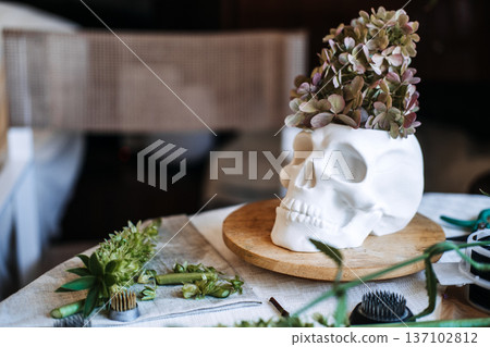White skull planter sits on wooden base with dried flowers on table. Custom home accents, edgy product design, unique craftsmanship. White skull planter sits on wooden base with dried flowers on table. Custom home accents, edgy product design, unique craftsmanship. 137102812
