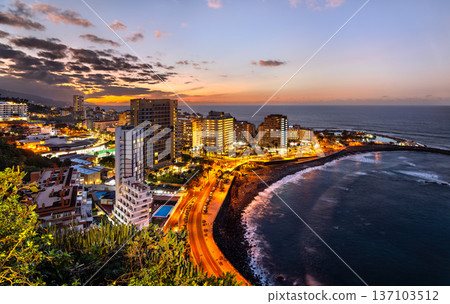 Scenic twilight view of the illuminated hotels, coastal road, and Lago Martianez municipal pool complex in the city of Puerto de la Cruz in Tenerife, Canary Islands, Spain. Scenic twilight view of the illuminated hotels, coastal road, and Lago Martianez municipal pool complex in the city of Puerto de la Cruz in Tenerife, Canary Islands, Spain. 137103512