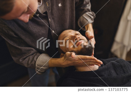 Barber massaging beard and neck of male client after shaving in barbershop. Relaxing grooming service, men self care, comfort and classic barber ritual concept. Barber massaging beard and neck of male client after shaving in barbershop. Relaxing grooming service, men self care, comfort and classic barber ritual concept. 137103775