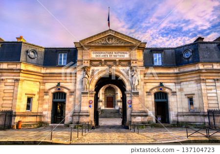 Scenic view of the historic classical stone facade, statues, and archway entrance of the Conservatoire National des Arts et Metiers building under a colorful sky in Paris, France. Scenic view of the historic classical stone facade, statues, and archway entrance of the Conservatoire National des Arts et Metiers building under a colorful sky in Paris, France. 137104723